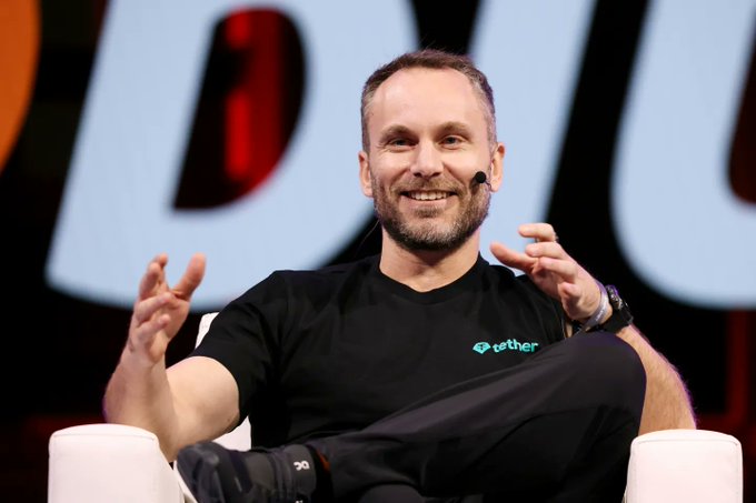 The image features a man sitting in a chair, smiling at the camera while wearing a black t-shirt with "Tether" written on it. The background shows an orange sign that reads "DUJI", which could be related to the event or company he is associated with. The text "Series C funding round for NEURA Robotics It is set to be one of the largest robotics funding rounds ever The funding will aid NEURA to Power the Financial and Intelligence Layer of the Robotics Era" suggests that the man might be involved in a project related to NEURA Robotics, possibly discussing their Series C funding round.