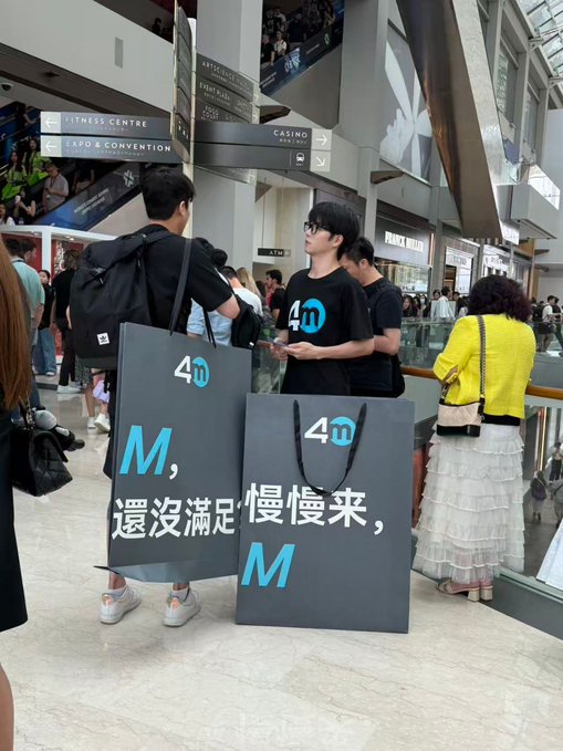 Two young men in black '4' logo t-shirts pose with large shopping bags reading '4M' and 'M，還沒滿足慢慢來，M' in a shopping mall.