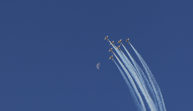 A flight of jets in formation leaving contrails across a deep blue sky near a crescent moon.