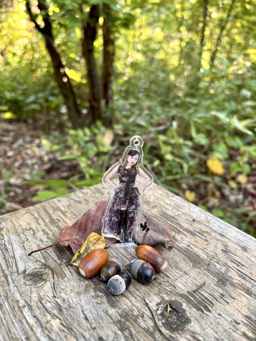 A clear acrylic charm depicting a woman in a black dress placed on a weathered wooden surface in a forest setting, surrounded by autumn acorns and leaves.