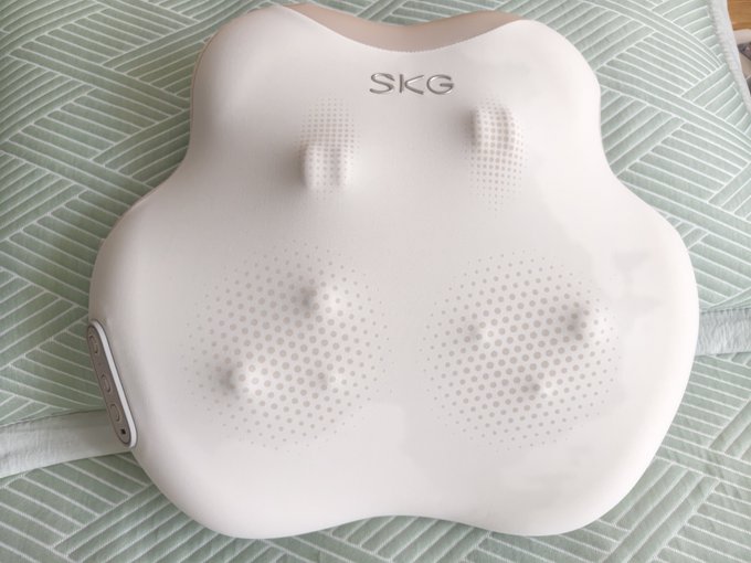 A white SKG massage device with a flower/cloud-shaped design featuring nodes for massage, resting on a green and white striped fabric surface.