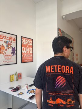 A person with glasses stands facing a white display table of small toys and cards, wearing a black t-shirt with a 'METEORA' graphic from the 'Rebuilding the City ofrobos of the Future' game, showing dates 07/2025, 17/0000, and 1201/2025. Framed 'BUY, SELL AND TRADE' and 'RADOST COFFEE CARDS' posters hang on the wall behind.