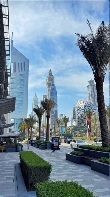 The image shows a modern cityscape featuring a clock tower, sleek skyscrapers, and palm trees lining a pedestrian walkway under a partly cloudy sky. The post "GM" likely refers to a "Good Morning" greeting, with the bright, sunny urban morning scene serving as a fitting backdrop.