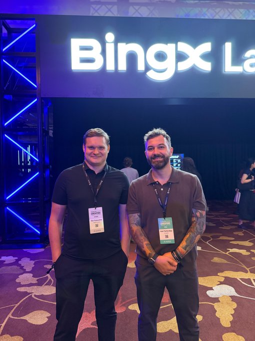 Two smiling men pose for a photo at an event with a large illuminated "Bingx" sign in the background; both wear lanyards labeled "VIP" and "BUYEREXPRESS".