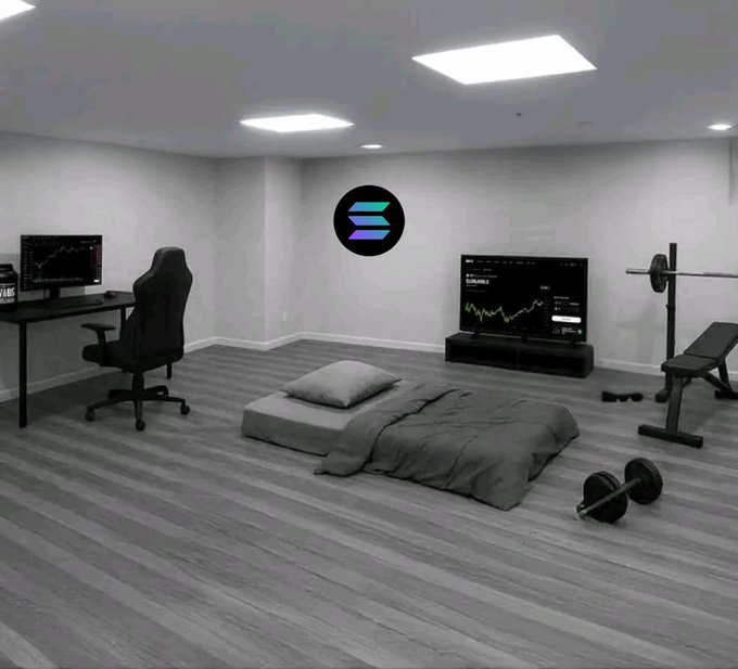 A dimly lit bedroom with a minimalist setup featuring a low mattress on the floor, a gaming chair at a computer desk displaying a chart, a TV showing a trading chart, and home gym equipment including a weight bench and barbell, with a Solana logo on the wall.