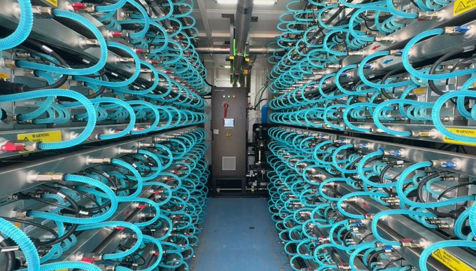 An interior view of a large data center server room with rows of rack-mounted servers on both sides, featuring numerous teal/turquoise Ethernet cables connecting the equipment.