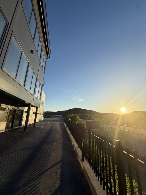 The image features an outdoor scene with a building and a fence. The sun is setting behind the building, casting long shadows across the area. A person can be seen standing near the fence, possibly enjoying the view or attending to their property.