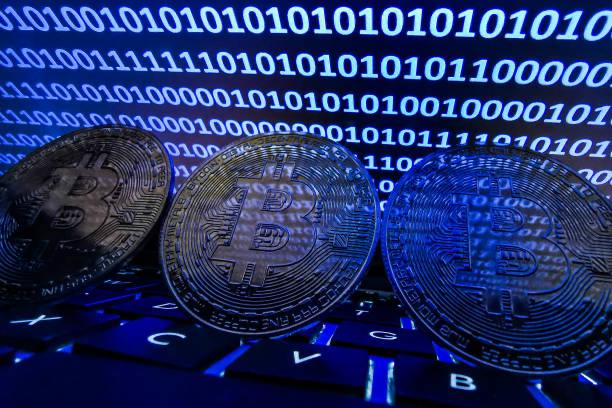 Three physical Bitcoin tokens rest on a backlit keyboard in front of a blue screen filled with binary code. The image visually reinforces the post's warning by symbolizing the digital cryptocurrency assets that Coldcard users must urgently relocate to a new wallet address following the identified security flaw.
