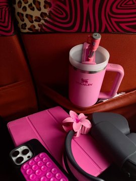 A pink Stanley insulated tumbler with a straw sits in a cup holder alongside a pink phone case with bubble keys and a black bag, against a leopard and zebra print background.