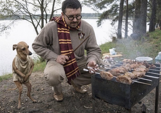 A man wearing round glasses, a Gryffindor scarf, and a Hogwarts sweater squats by a charcoal grill, cooking skewers of meat outdoors by a lake while a thin dog sits beside him.