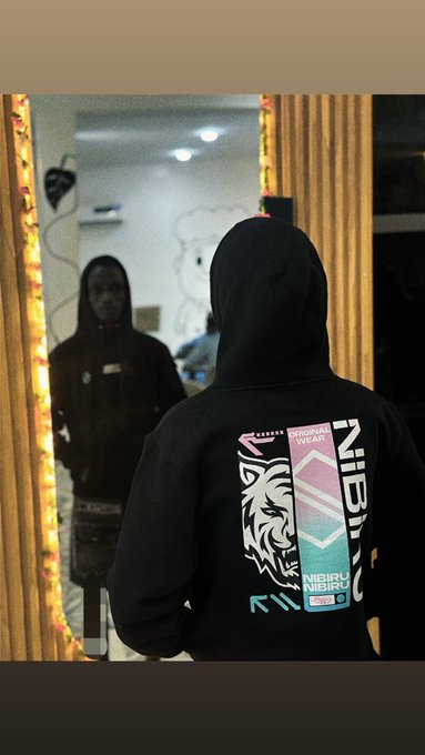 A person wearing a black hoodie with a 'NIBIRU NISEKU ORIGINAL WEAR' graphic on the back, standing in front of a mirror reflecting their face, with orange decorative lights framing the mirror.