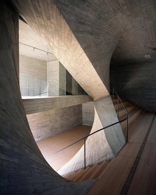 An interior architectural shot showcasing a striking concrete staircase with curving, sculpted walls and wood flooring, likely from a modern museum or cultural building.