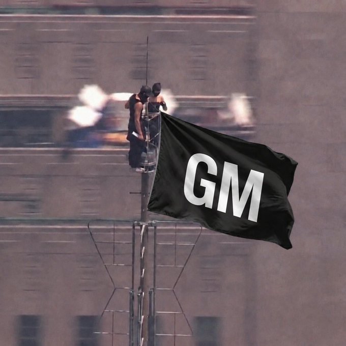 Two masked individuals on a tall building structure raise a black flag with bold white 'GM' lettering against an urban backdrop.