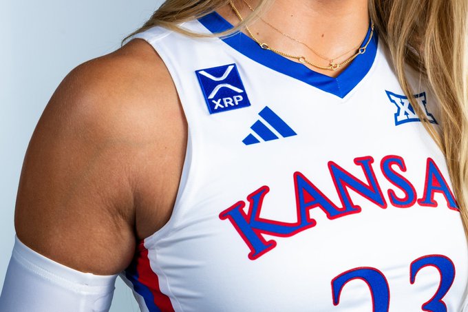 A close-up shot of a white Kansas basketball jersey featuring a blue XRP sponsor patch on the chest alongside the Adidas logo, with the number "23" partially visible and a Big 12 conference emblem. The image showcases the partnership mentioned in the post, demonstrating how the XRP logo is being displayed on collegiate athletic apparel as part of a strategy to boost brand recognition against competitors like BTC.