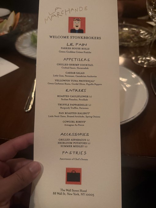A hand holding a custom dinner menu titled 'MARCHANDE - WELCOME STONKBROKERS' from The Wall Street Hotel, featuring courses like Le Pain, Appetizers, Entrees, and Pastries.