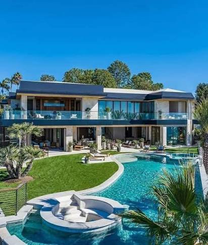 A luxurious modern two-story home with expansive glass windows overlooks a stunning curved swimming pool with an attached hot tub. The property features a manicured green lawn, tropical landscaping with palm trees, and outdoor lounge areas under a clear blue sky.