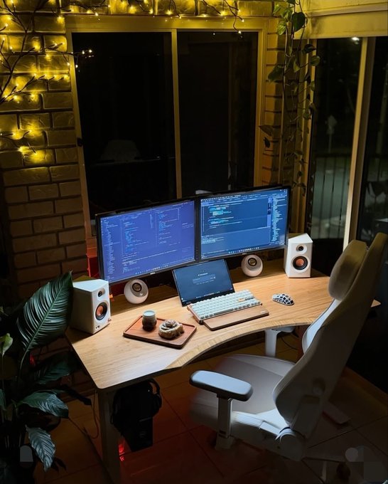 A home office setup at night with a curved desk holding two monitors displaying code, a laptop, speakers, and an ergonomic chair, surrounded by warm string lights.