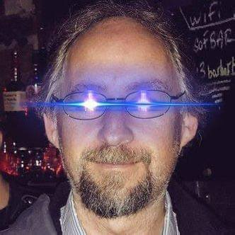 A close-up portrait of a man with glasses and a beard, wearing a striped shirt, with a distinctive blue light reflection across his lenses.