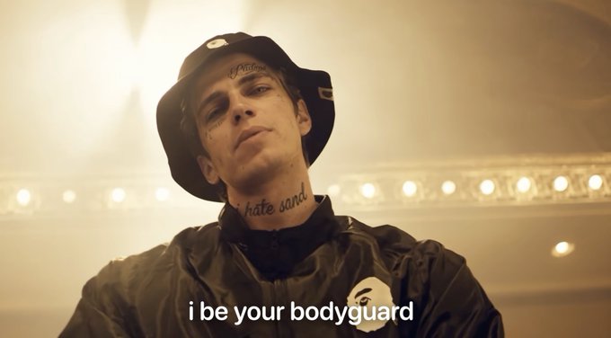 A music video still of Justin Bieber wearing a black bucket hat and jacket, with visible face/neck tattoos reading 'Peach' and 'hate sand,' with subtitles reading 'i be your bodyguard'.