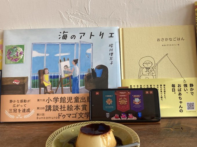 A cozy Japanese setting with two picture books displayed behind a smartphone playing a colorful card game, with a caramel pudding dessert in the foreground on a wooden surface.