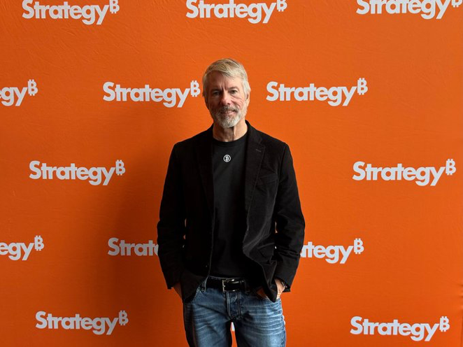 A man with gray hair and a beard, wearing a black blazer and dark shirt, stands in front of an orange backdrop featuring the "Strategy" logo. The image appears to be from a Strategy company event, aligning with the post about Michael Saylor's Strategy selling 1,691 $BTC ($108 million) and reducing its total holdings to 840,447 BTC.