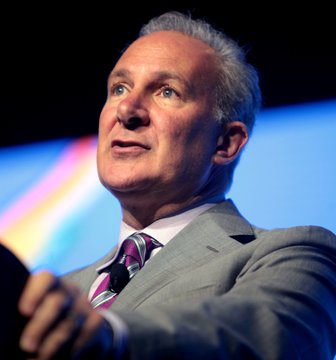 A close-up photograph captures a gray-haired man in a gray suit and striped tie, appearing to speak at an event with a colorful backdrop.