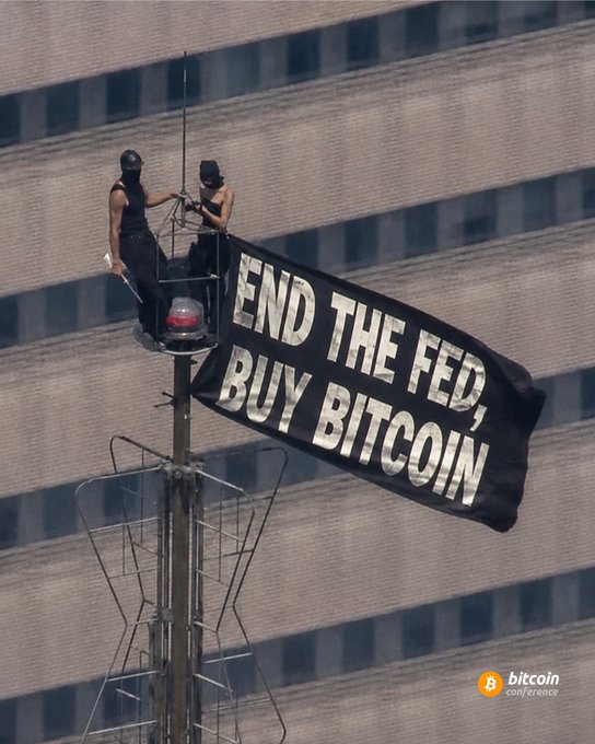 Two masked activists unfurl a banner reading "END THE FED, BUY BITCOIN" from atop a pole in front of a skyscraper, visually reinforcing the post's call to abandon the Federal Reserve in favor of Bitcoin. The Bitcoin Conference logo appears in the bottom right corner, linking the protest to the cryptocurrency community's anti-central bank sentiment.