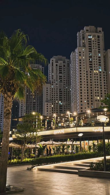 A nighttime cityscape features illuminated high-rise towers behind an outdoor restaurant with visible "AWAN" and Arabic signage, with palm trees and lampposts framing a paved plaza.