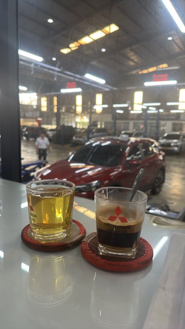 Two drinks sit on red coasters on a white table: a glass of tea and a small cup of espresso with a Mitsubishi logo, overlooking a red car parked inside a brightly lit automotive service center.