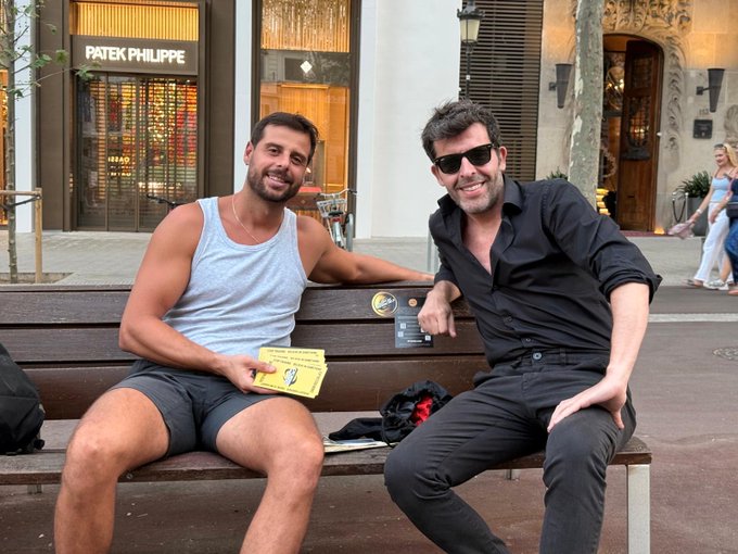 Two men smiling while sitting on a bench on a city street, with a 'PATEK PHILIPPE' storefront visible in the background.
