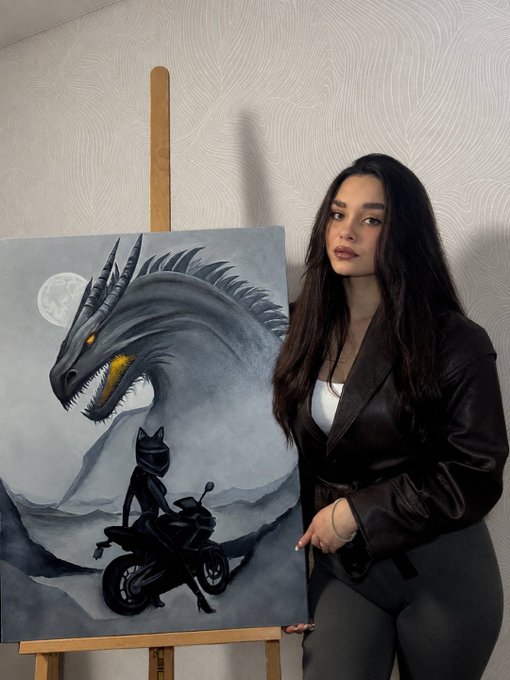A woman with long dark hair in a black leather outfit stands beside an easel displaying a painting of a black dragon-like creature and a helmeted figure on a motorcycle.