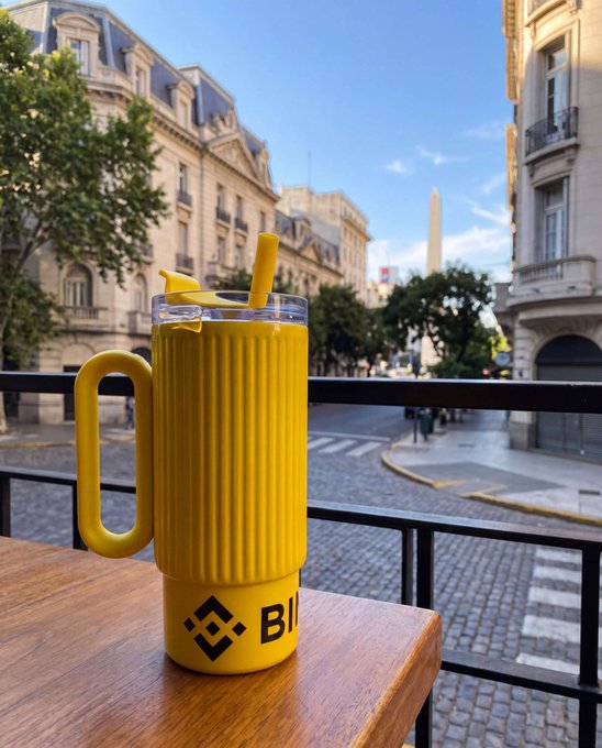 A yellow mug with the Binance logo sits on a wooden table, filled with a yellow drink and lemon garnish, overlooking a street in Paris with an obelisk in the background.