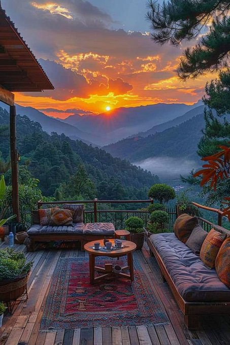 A wooden outdoor deck with cushioned seating and a coffee table overlooks a lush mountainous valley at sunset, with warm orange light filtering through clouds.