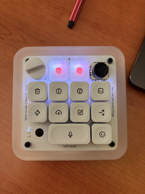 The image shows a custom "Work louder! | OpenAI 2026" macro pad with illuminated keys, including a spacebar labeled "Let's build" and the phrase "You can just build things," sitting on a wooden desk. The caption "Removing agent keys is bold" ties into the device's playful push-button interface, where one literally removes the "agent" key while the macro pad embodies a hands-on, keys-at-your-fingertips approach to AI agent work.