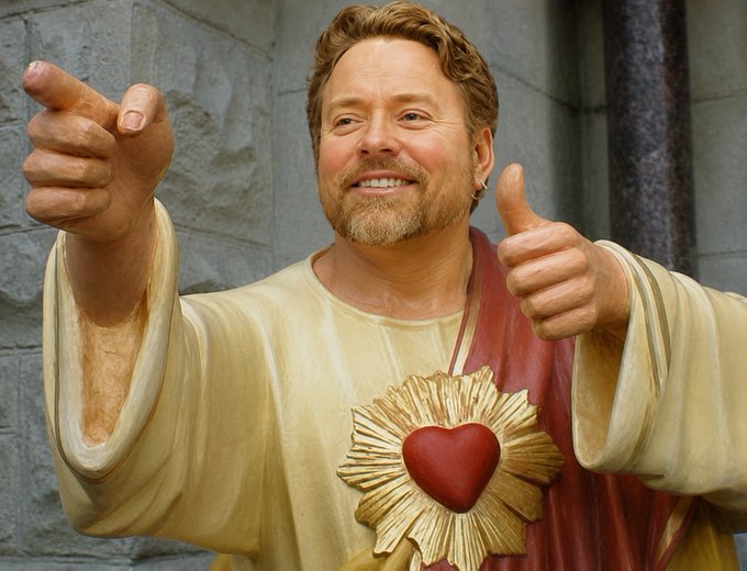A man with reddish-blonde hair and a goatee poses wearing a yellow and red robe with a Sacred Heart emblem, giving two thumbs up while smiling.