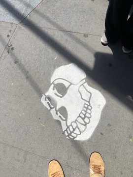A white skull stencil sprayed on a sidewalk, with two pairs of feet visible at the edges of the frame.