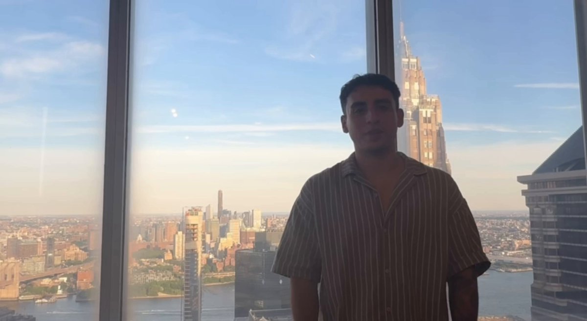 The image features a man standing next to a large window with a view of the city skyline outside. The man appears to be posing for a photo while looking directly at the camera. The backdrop includes a tall building, possibly the New York City skyline, which is visible through the glass window. The post seems to be promoting an event or gathering related to cryptocurrency and DeFi, as indicated by the text in the image.