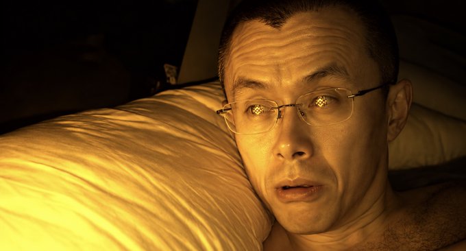 A close-up shows a man with short dark hair and glasses lying on a white pillow, his face illuminated by warm golden light in a dark room.