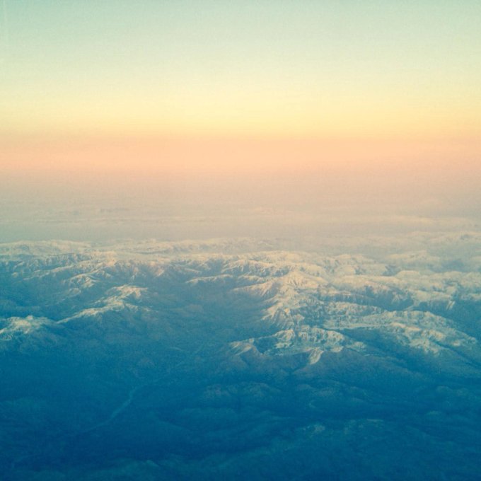 The image shows a beautiful view from an airplane window, with mountains covered in snow visible below. The sky above has a warm orange hue, creating a stunning contrast between the blue of the sky and the white snow-covered peaks. The photo is taken from an aerial perspective, providing a unique vantage point to appreciate the breathtaking scenery.