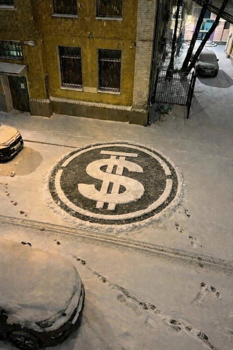 A large dollar sign symbol within a circle is drawn or stamped in the snow on a city street at night, with footprints and a parked car visible.