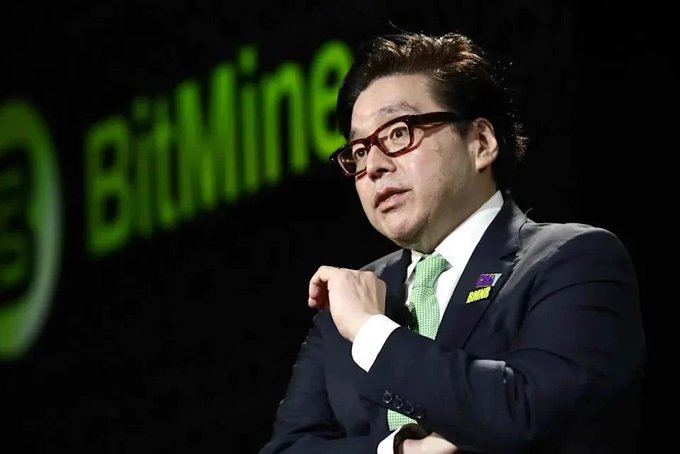 A man in a dark suit and green tie wearing glasses stands in front of a blurred 'BitMine' logo on a green background.