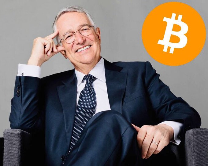 A confident, smiling older man in a navy suit and patterned tie sits in a gray chair beside a Bitcoin logo, visually pairing the financial figure referenced in the post—Mexican billionaire Ricardo Salinas—with his endorsement of Bitcoin as a hard asset resistant to fiat currency inflation.