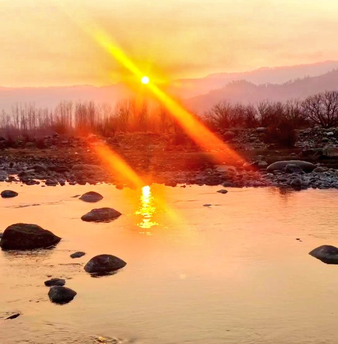 A sunset over a rocky river creates bright yellow and orange light rays reflecting off the water's surface.