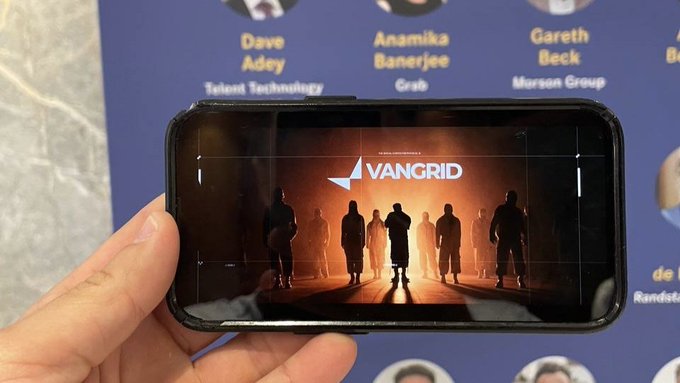 A hand holds a smartphone displaying a website or app for "VANGRID" featuring silhouettes of a team against a fiery orange background.