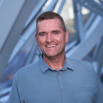 A smiling middle-aged man in a blue polo shirt stands in front of a modern geometric architectural background featuring angular blue structural beams. The image relates to the post as it likely depicts Dave Brown, the former AWS executive recently hired by Meta to lead their data-center buildout and Meta Compute cloud venture.