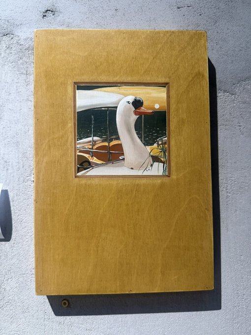 A gold-colored rectangular frame mounted on a white textured wall holds a small painting of a white duck with an orange beak.