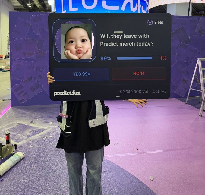 A person poses behind a large "predict.fun" prediction board at an event, with the question "Will they leave with Predict merch today?" showing 99% YES and 1% NO, $2,049,000 Vol for Oct 7-8, humorously holding up the board to match their own caption about leaving with @predictdotfun merch.