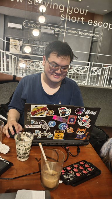 A man with glasses sits smiling at a laptop covered in colorful stickers (including "Linear," "Auto," and various meme graphics), with an iced coffee and strawberry-patterned phone case on the table before him. The caption's reference to "ibu-ibu" (Indonesian for "moms") and "Yunepto" appears to be teasing him about his crypto community involvement, likely related to the stickers visible on his work laptop.