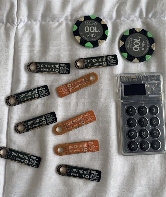 Several Opendime Bitcoin hardware wallets and two ARIA poker chips ($100) are arranged on a white cloth next to a Casio-style calculator.