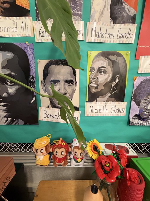 A classroom display features student-made portraits of historical figures labeled "Muhammad Ali", "Mahatma Gandhi", "Barack", and "Michelle Obama", with small plush toys and artificial flowers arranged below.